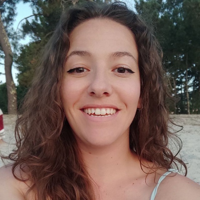Woman with curly and loose hair, smiling outdoors, with trees and sand in the background, under natural light.